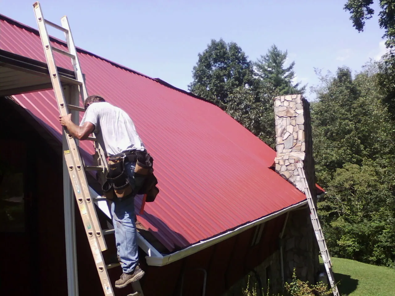 Roofer on a ladder beside a red metal roof and stone chimney — Soffit & Fascia Repair by Roofing Brothers in Parlier